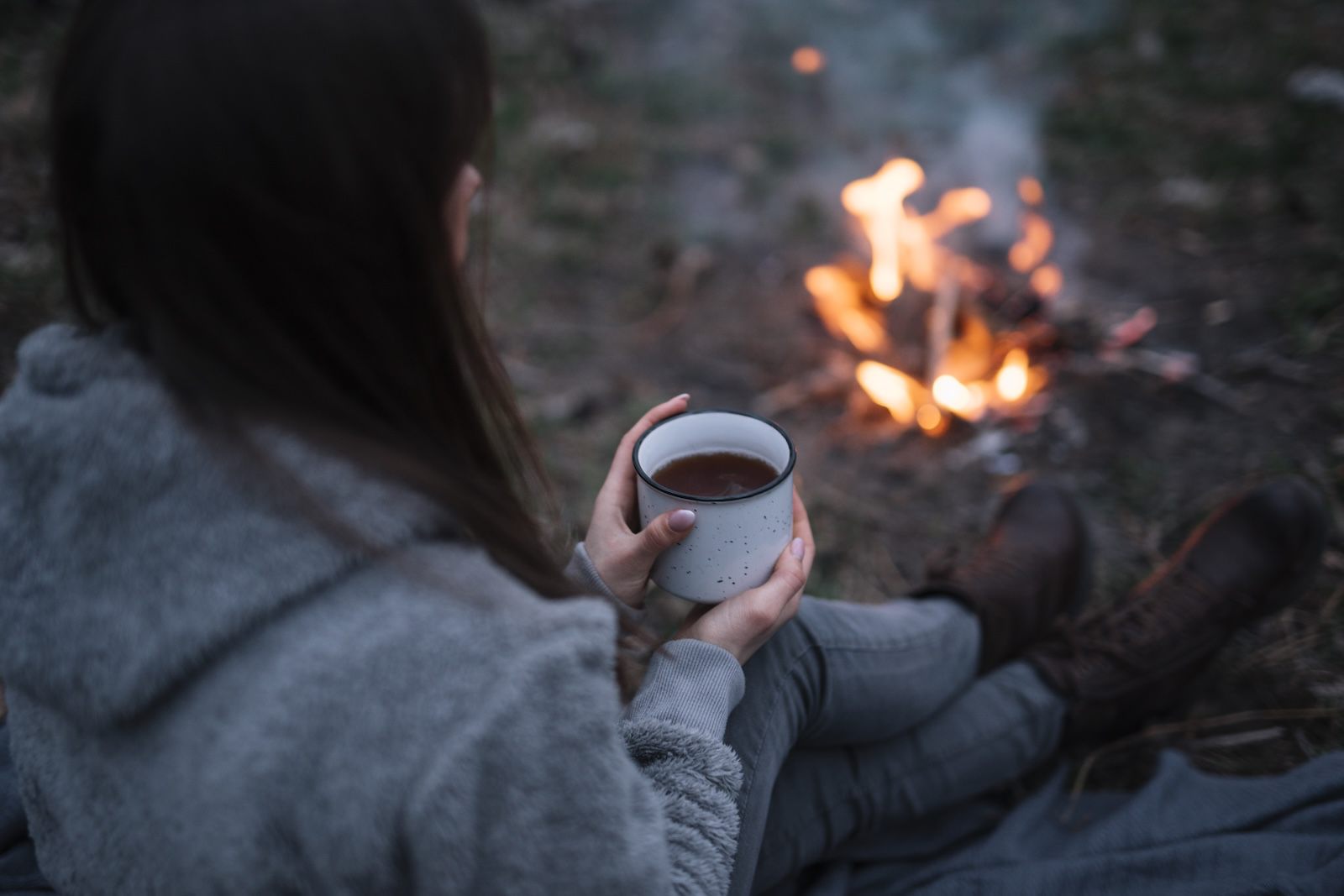 Person sitzt am Lagerfeuer und hält warme Tasse im Wald