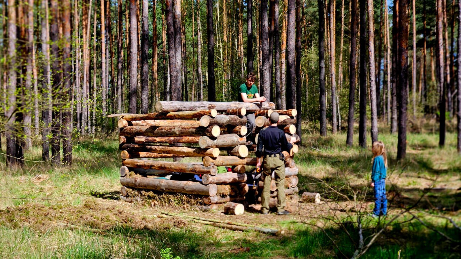 Selbstgebautes Blockhüttenhaus im Wald bei dem zwei Jugendliche und ein Kind stehen